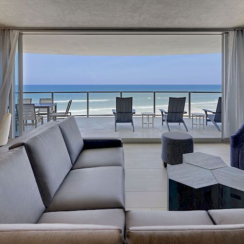 A modern living room with a large sofa, chair, and table, leading to a balcony with a stunning ocean view in the background.