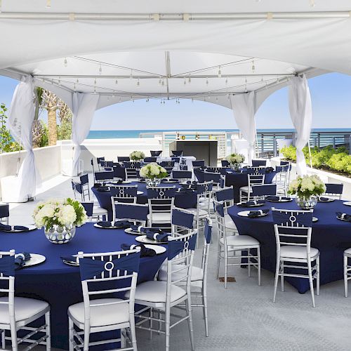 The image shows an outdoor event setup with round tables covered in navy blue tablecloths and white chairs, under a white canopy near the beach.