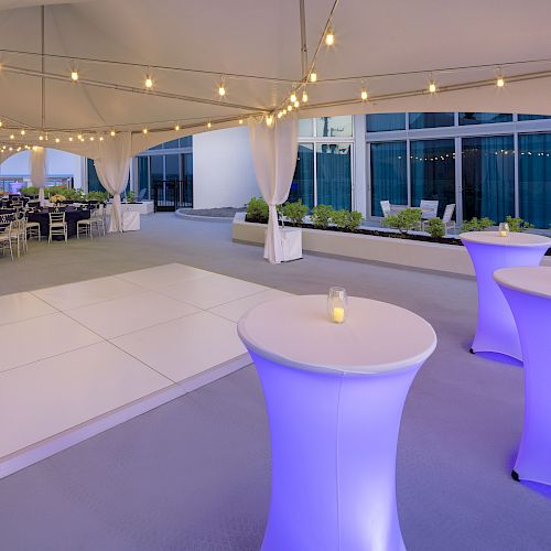 An outdoor event setup features a dance floor, tall illuminated tables with candles, draped tent, string lights, and tables with chairs in the background.