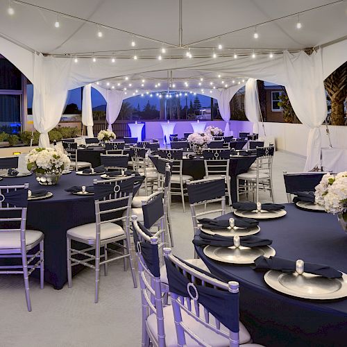 An elegantly decorated outdoor event setup with round tables covered in navy blue tablecloths, silver chairs, floral centerpieces, string lights, and a canopy.