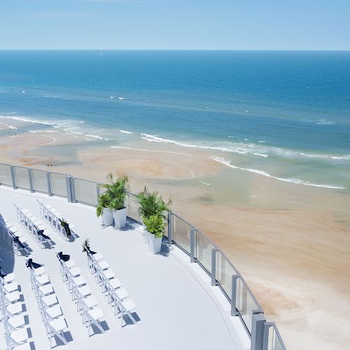 A beautiful coastal view with a modern terrace setup that includes white chairs and potted plants, overlooking a sandy beach and the vast ocean.