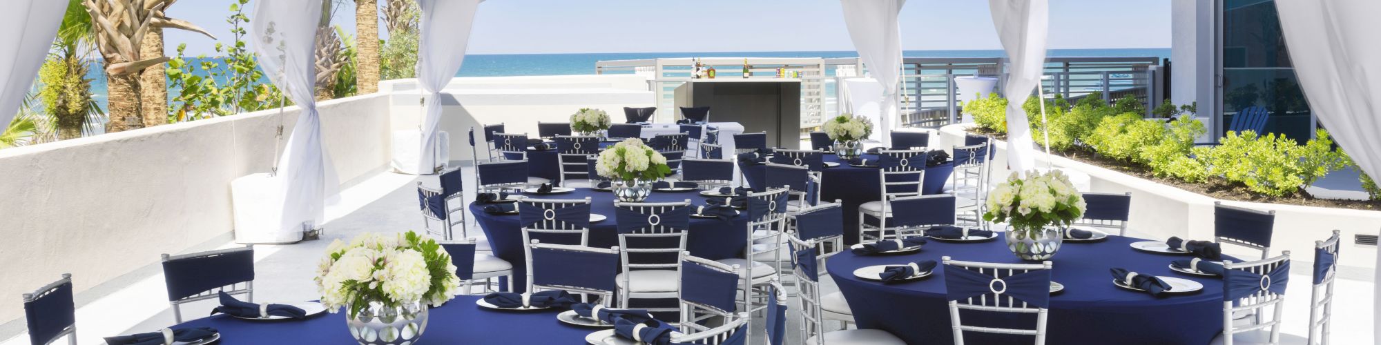 The image shows an outdoor event setup with round tables covered in navy tablecloths, white chairs, floral centerpieces, and a white canopy.