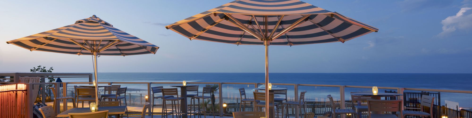 This image shows a seaside patio with tables and chairs under large striped umbrellas, set against a backdrop of the ocean at sunset.