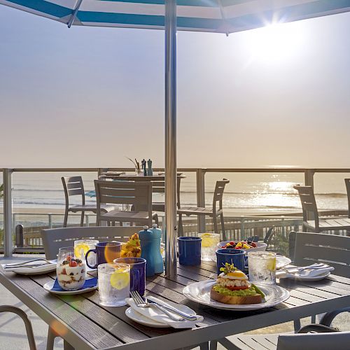 An outdoor dining setup with an umbrella, food, drinks, and tableware on a wooden table. Background shows a sunny beach and blue sky.