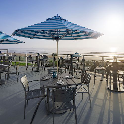 Outdoor seating area with tables, chairs, and umbrellas overlooking a serene beach at sunrise or sunset.