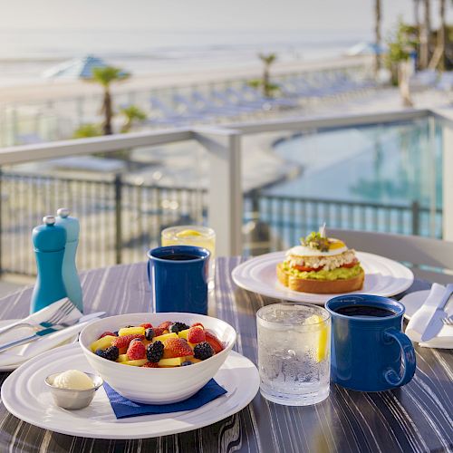 A breakfast setup on a patio includes a fruit bowl, toast with avocado, mugs, glasses of juice, and utensils, with a pool and beach view.