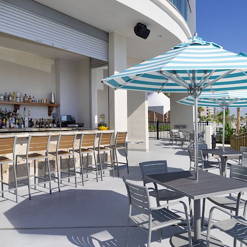 An outdoor bar with stools, tables, chairs, and umbrella shades. It is a sunny day and the bar is stocked with various bottles and glasses.