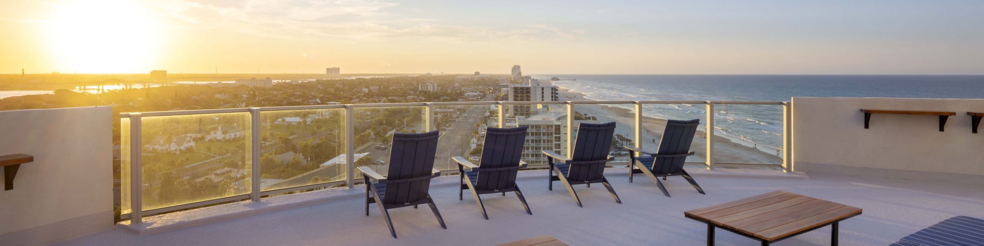 A rooftop terrace with lounge chairs and tables overlooks an ocean and a city at sunset, providing a serene and scenic view.