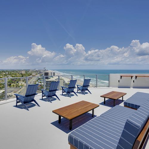 This image shows a rooftop lounge area with blue chairs, tables, and a long sofa overlooking a scenic ocean view and a city skyline.