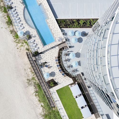 An aerial view of a beachfront hotel, showing a pool, lounge area, and balconies with chairs, adjacent to a sandy beach and parking lot.