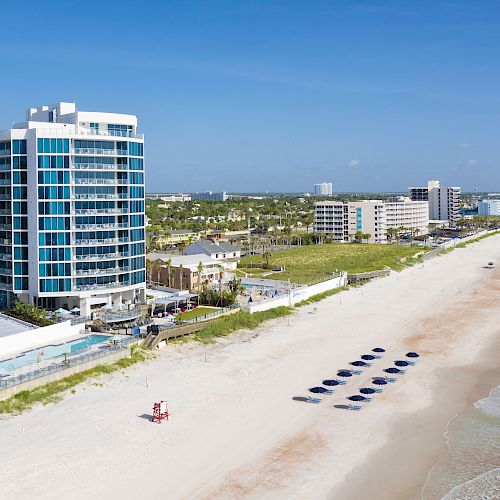 A coastal scene featuring a tall hotel with an adjacent pool, a sandy beach with lined-up umbrellas, and waves crashing on the shore.