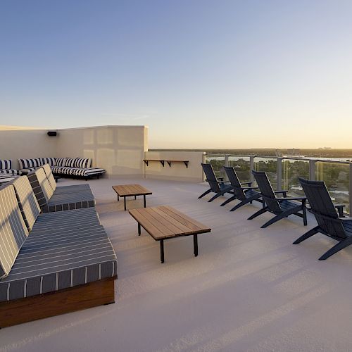 The image depicts a rooftop terrace with lounge chairs, benches, and tables, overlooking a scenic view at sunset, with glass railings along the edge.