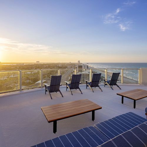 A rooftop terrace with lounge chairs and wooden tables overlooks the cityscape and ocean during sunset.