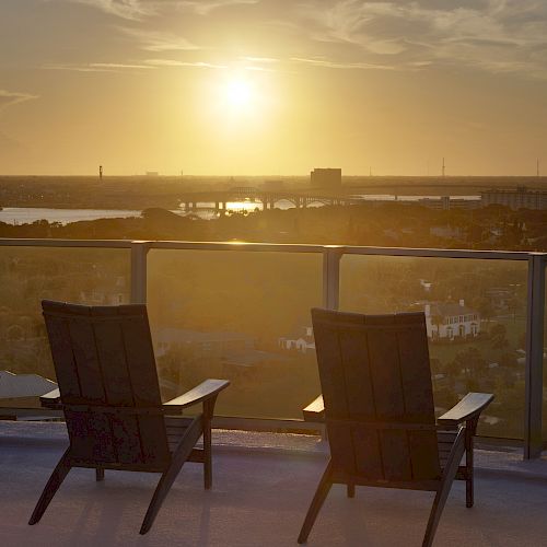 The image shows a sunset view from a rooftop terrace with several empty lounge chairs facing a scenic landscape.