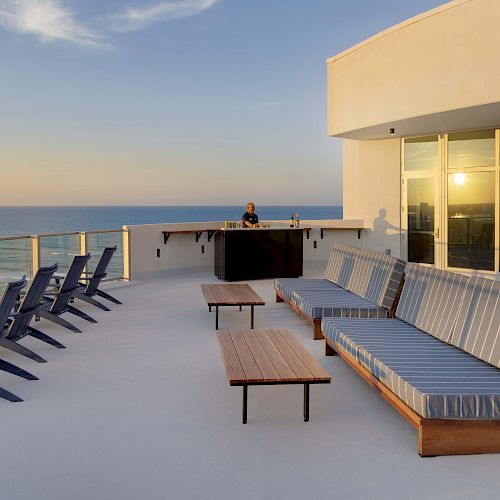 A rooftop patio with lounge chairs, sofas, and tables facing an ocean view. A person stands behind a bar counter in the background.