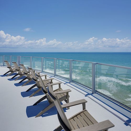 Wooden deck chairs line a balcony overlooking the ocean, with clear glass railings and a bright, blue sky in the background.