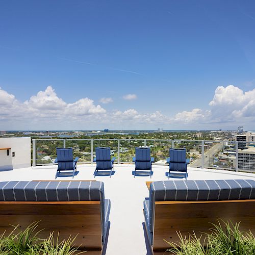 This image shows a rooftop terrace with blue lounge chairs, overlooking a cityscape and ocean horizon under a blue sky with scattered clouds.