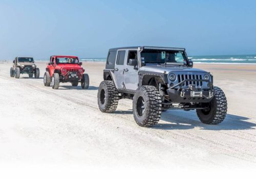 A trio of lifted off-road SUVs drives on a sunny sandy beach with ocean waves in the background.