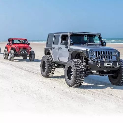 A trio of lifted off-road SUVs drives on a sunny sandy beach with ocean waves in the background.