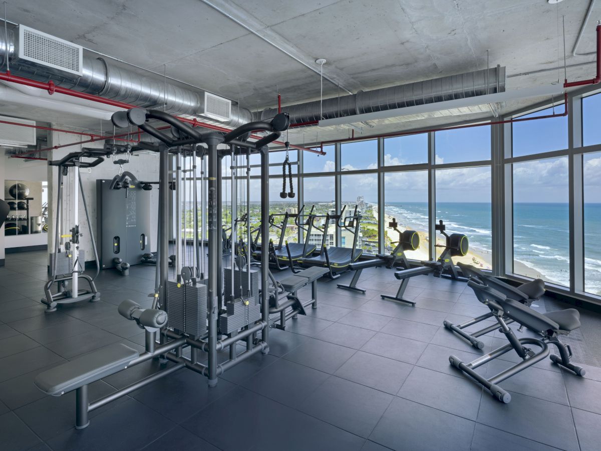 A modern gym with various workout equipment, large windows offering a beach view, and an industrial ceiling design.