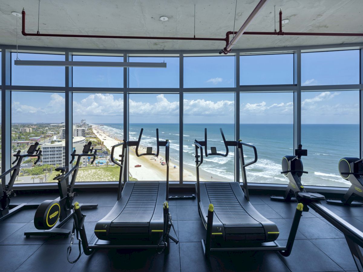 A gym with treadmills and exercise bikes, featuring large windows displaying a scenic ocean and beach view on a sunny day.