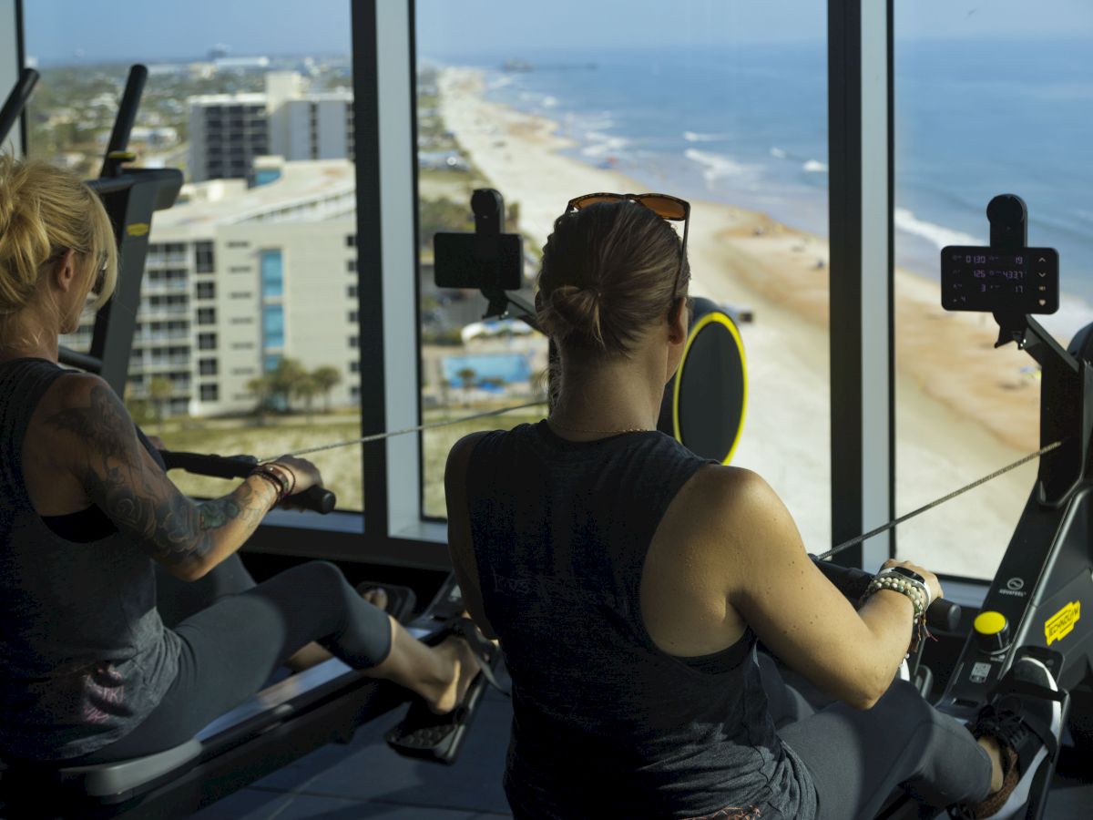 Two people rowing on exercise machines in a gym with large windows offering a scenic view of the beach and ocean outside.