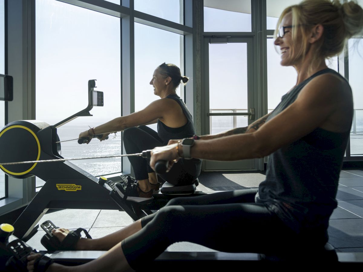 Two people are exercising on rowing machines in a room with large windows that offer a scenic view of the outdoors.