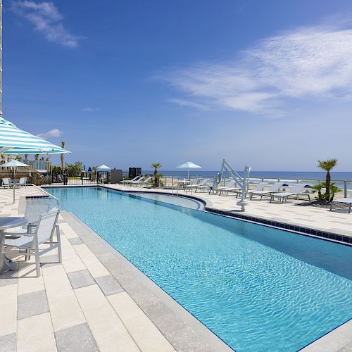 An outdoor pool with lounge chairs, tables with umbrellas, and a view of the ocean. The sky is clear with a few clouds.