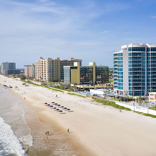 A beach with waves in the foreground, buildings in the background, and a few people walking on the sand under a clear blue sky.