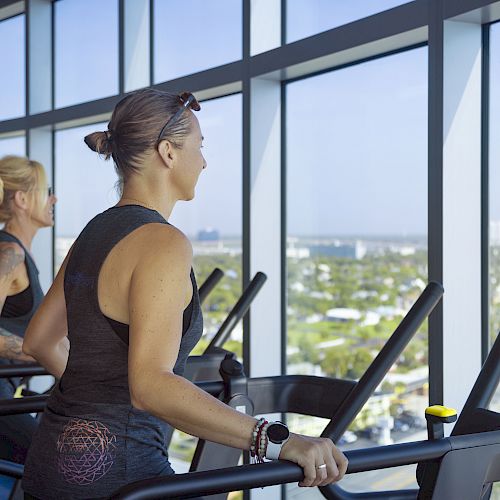 Two individuals are exercising on treadmills in a gym with large windows offering a view of a cityscape and clear sky in the background.