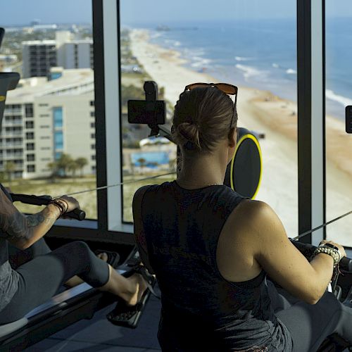 Two individuals are using rowing machines in a high-rise gym with large windows offering a scenic view of a beach and ocean in the background.