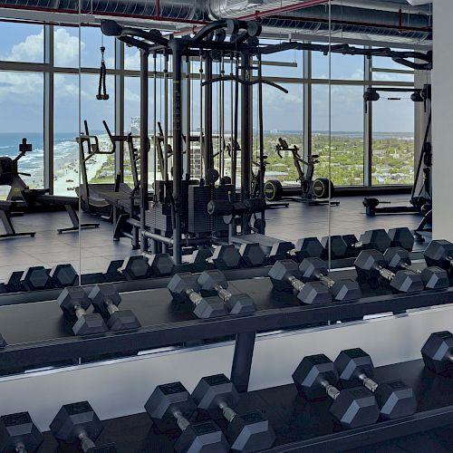 A gym with rows of dumbbells, weight machines, and a panoramic window offering an ocean view, with cardio equipment visible in the background.