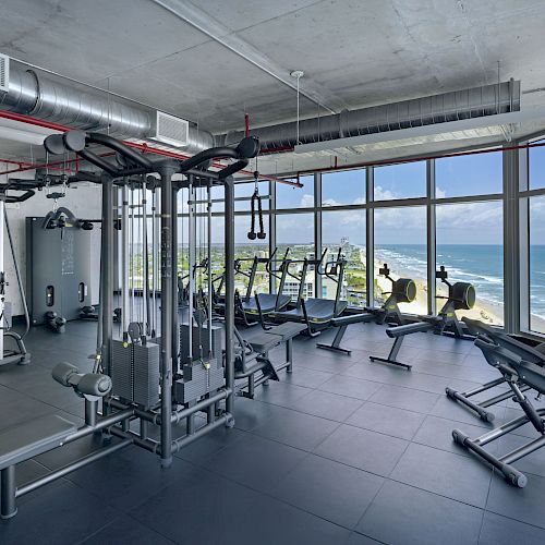 A modern gym with various exercise equipment is shown, featuring large windows with a view of the ocean and coastline under a blue sky.