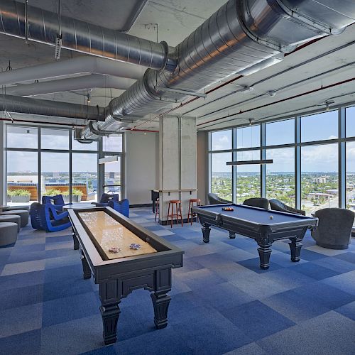 A modern game room with a pool table, shuffleboard, seating, floor-to-ceiling windows, and city views in the background.