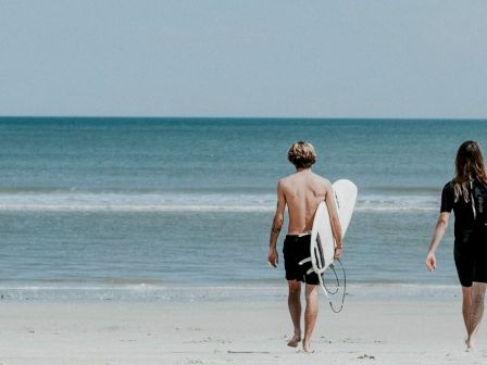 Two people walking towards the ocean, each carrying a surfboard, with a peaceful beach and calm sea in the background.