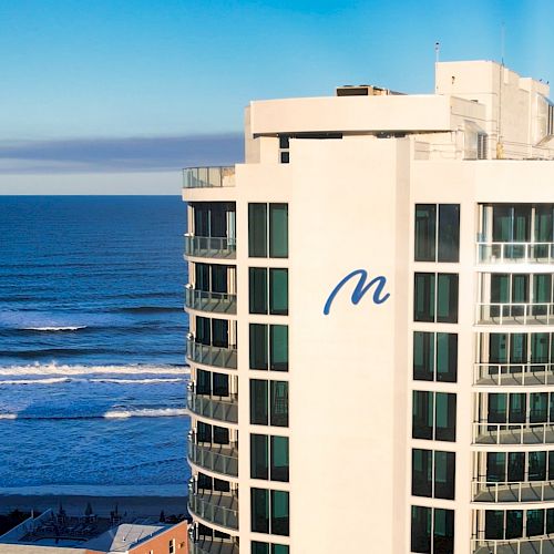 A tall building with a blue "M" on it is situated near an ocean with waves and a clear blue sky in the background.