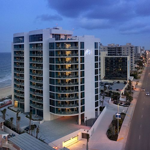 A tall, modern building by the beach at dusk, with a well-lit road and more buildings in the background, under a partly cloudy sky.