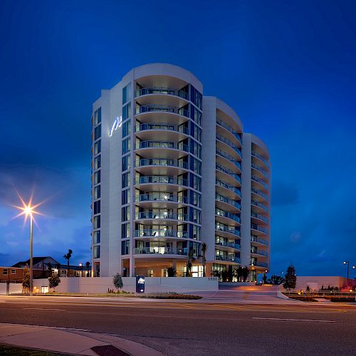 A modern multi-story building is lit up at night under a deep blue sky, surrounded by streets and a few trees.