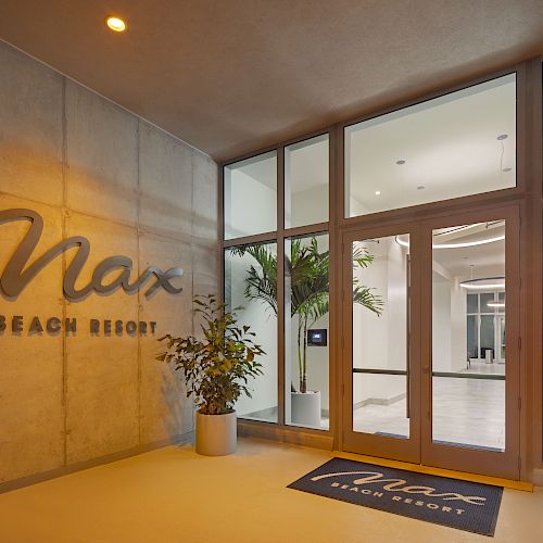 Entrance to "Max Beach Resort" with logo on wall, potted plants, glass doors, and a welcome mat. Modern, well-lit interior visible.