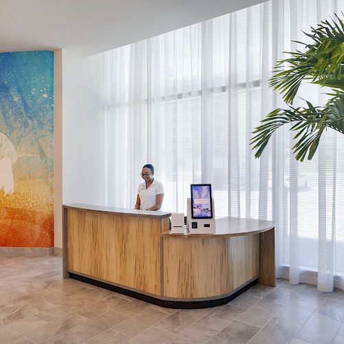 A person at a wooden reception desk, with a colorful mural on the left and a large plant on the right, in a bright, modern lobby.