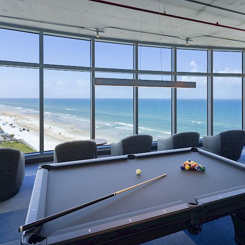 A pool table in a room with large windows offering a scenic view of the beach and ocean, surrounded by chairs and a stool.