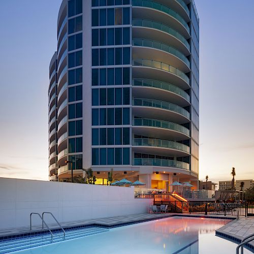 The image depicts a modern high-rise building next to a swimming pool at dusk, with the pool water reflecting the surrounding lights.