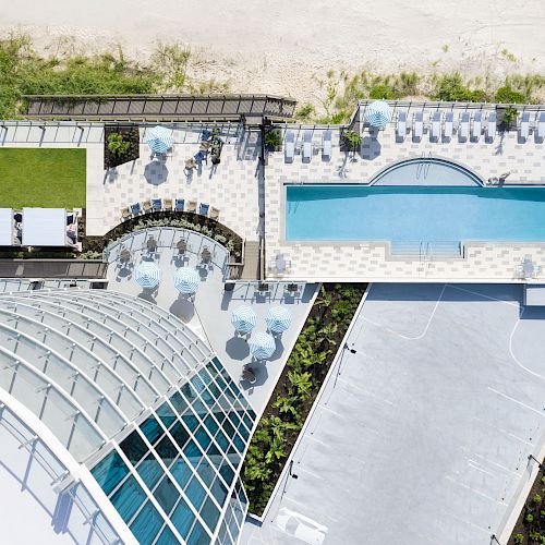 Aerial view of a resort featuring a swimming pool, sun loungers, green lawn area, and a glass-roofed structure near a beach.