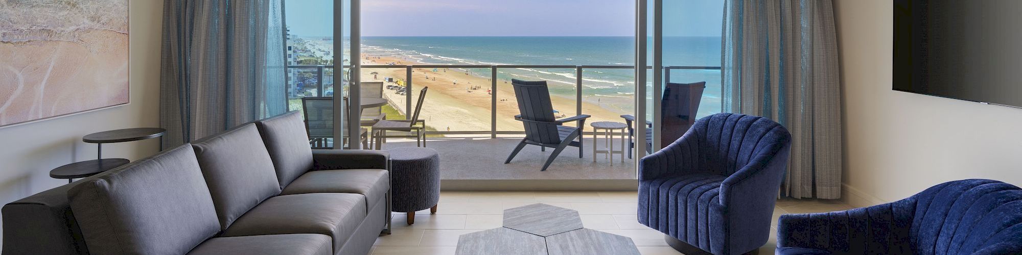 A modern living room with a beach view, featuring a sofa, two armchairs, a TV, and a unique coffee table, opens onto a balcony with seating.