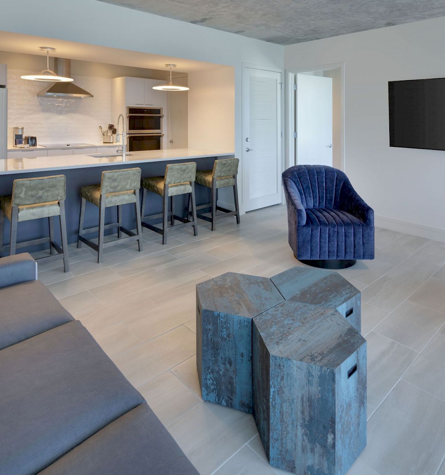 A modern living room with blue furniture, a wall-mounted TV, and a kitchen area with bar stools and stainless steel appliances in the background.