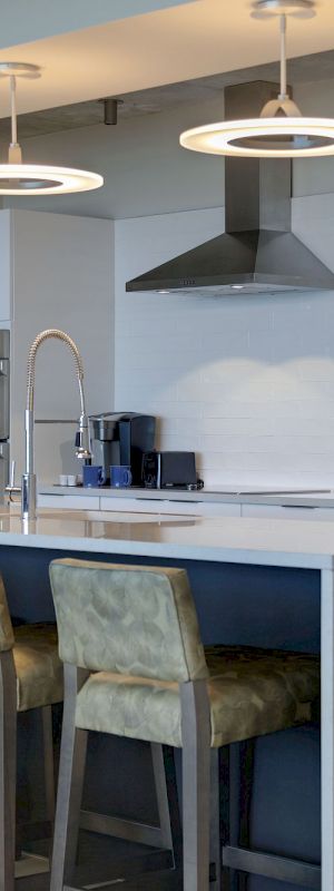 A modern kitchen with an island, bar stools, pendant lights, stainless steel appliances, and white cabinets.