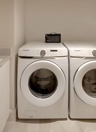 The image shows a laundry room with a washing machine and a dryer placed side by side, a sink, and a shelf with detergent.