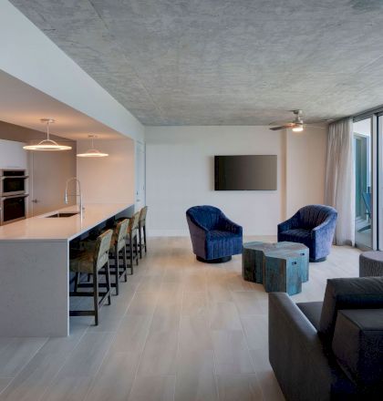 A modern living space features a kitchen and island, seating area with blue chairs, a TV, and an ocean view from large glass windows leading to a balcony.