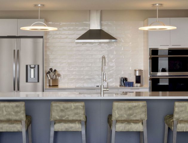 A modern kitchen with stainless steel appliances, pendant lights above the counter, and four upholstered bar stools at the island.