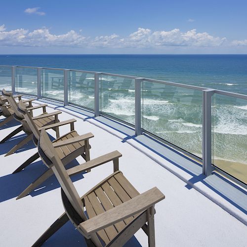 Multiple wooden lounge chairs are arranged on a balcony with a glass railing, overlooking a scenic ocean view under a clear blue sky.